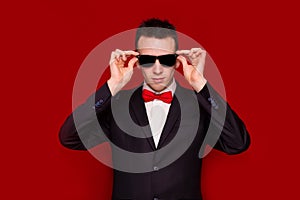 Portrait of serious handsome stylish man in elegant black suit and sunglasses on red background