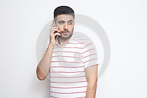 Portrait of serious handsome young adult man wearing T-shirt standing, using smartphone, making call and talking with friend
