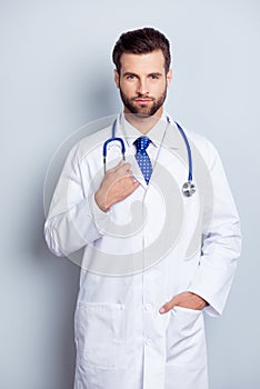 Portrait of serious handsome young male doctor holding hand in pocket standing on gray background