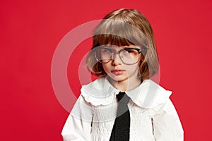 Portrait of serious little school girl in white blouse with tie and big glasses, looking seriously against red studio