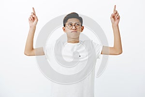 Portrait of serious-looking charming young chinese male student in white t-shirt raising hands, pointing and looking up