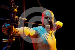 Portrait of serious man, archer aiming with bow on target against black studio background in neon light. Archery sport
