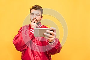 Portrait of a serious man with a beard, wearing a red jacket, stands with a smartphone in his hand on a yellow background and