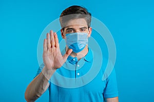 Portrait of serious man in professional medical mask showing rejecting gesture by stop palm sign. Guy isolated on blue