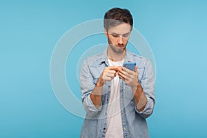 Portrait of serious man in worker denim shirt texting message in social media on cell phone, using mobile network