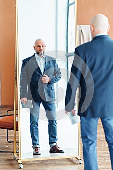 Portrait of serious middle-aged bald man with gray beard wearing blue suit jacket, holding notebook, looking at mirror.