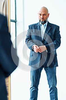 Portrait of serious middle-aged bald man with gray beard wearing blue suit jacket, looking gravely at mirror reflection. photo