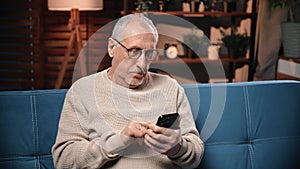 Portrait of serious old man surfing internet by smartphone in home, tapping and swiping on display