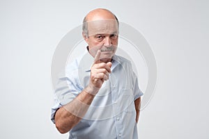 Portrait of serious senior man with warning finger and blue shirt shirt against light gray background.