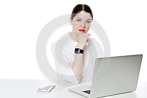 Portrait of serious successful brunette businesswoman in white shirt sitting with laptop, touhing her chin and looking at camera