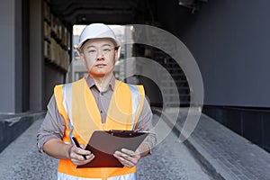 Portrait of serious and successful young Asian man in hard hat and vest standing outside building with folder and pen in