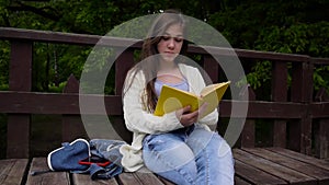 Portrait of serious teenage girl reading book and turning page leaning sitting on the bench in forest in spring