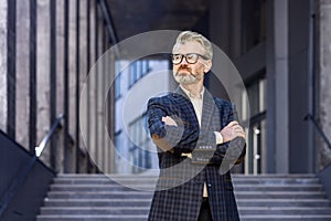 Portrait of serious thinking mature businessman, senior gray haired man in business suit outside office building