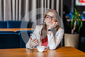 Portrait of serious thoughtful beautiful stylish brunette young woman in glasses sitting, holding her mobile smart phone and