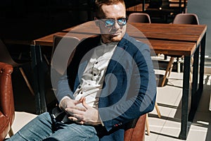 Portrait of serious thoughtful young man in sunglasses wearing stylish suit wear sitting in armchair, looking away out