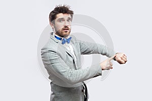 Portrait of serious worried handsome bearded man in casual grey suit and blue bow tie standing with watch hand gesture and looking