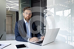 Portrait of a serious young businessman sitting in the office at a desk, working on a laptop and with documents, looking