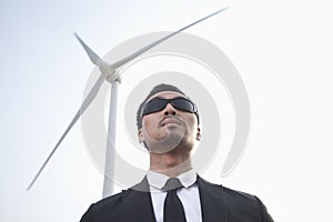 Portrait of serious young businessman in sunglasses standing by a wind turbine