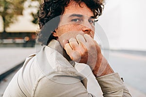 Portrait of serious young man with curly hair looking at the camera, sitting outside. Portrait of handsome male with curly hair