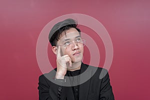 Portrait of a serious young man with glasses deep in thought, pointing at his head while looking up. Isolated on a red background