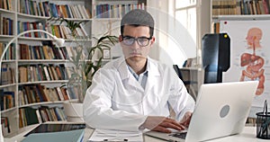 Portrait of serious young man looking at laptop screen and than to camera while sitting in medical office. Crop view of