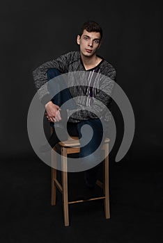 Portrait of serious young man is sitting on chair with one leg on it on black background