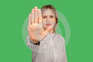 Portrait of serious young woman in classic striped blouse showing stop gesture to camera, warning with raised palm