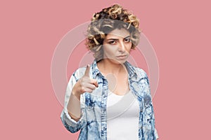 Portrait of serious young woman with curly hairstyle in casual blue shirt standing, looking at camera with warning hand