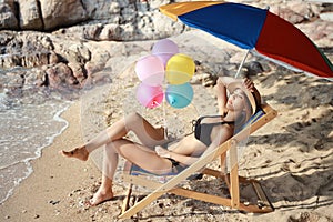 Portrait of sexy asian young girl in bikini and hat sitting on deck chair at tropical beach and colorful balloons with beauty face