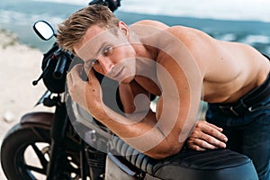 Portrait of sexy athletic man with naked torso on custom motorbike with ocean at background