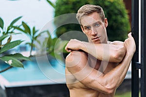 Portrait of sexy athletic man in white pants with naked torso resting in tropical garden