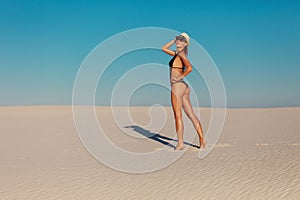 Portrait of sexy beautiful tanned model woman posing in fashion black swimwear bikini, fedora hat and sunglasses sand desert beach