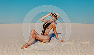 Portrait of sexy beautiful tanned model woman posing in fashion black swimwear bikini, fedora hat and sunglasses sand desert beach