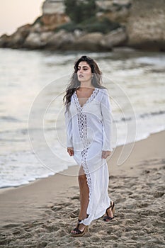 Portrait of a sexy female wearing a long white dress walking on the seashore of the beach