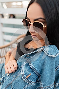 Portrait of a sexy pretty young woman with beautiful black hair in trendy sunglasses in a black t-shirt in a stylish denim jacket