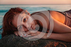 Portrait of a sexy tanned woman in lingerie, posing lying on her stomach, on the coastal rocks. Sea and sunset in the background.