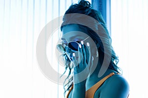 Portrait of a sexy young woman with curly hair in sunglasses with a stylish manicure in a trendy yellow swimsuit in a solarium.