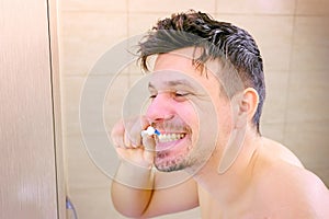 Shaggy sleepy young man intensively cleaning teeth in bathroom, side view. photo