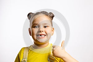 Portrait of shining little girl showing missing front baby tooth and thumb up in yellow t-shirt on white background