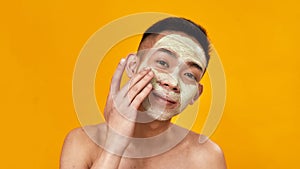 Portrait of shirtless young asian man applying green mask on his face isolated over yellow background. Beauty, skincare