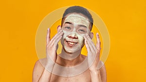 Portrait of shirtless young asian man smiling at camera while applying green mask on his face isolated over yellow