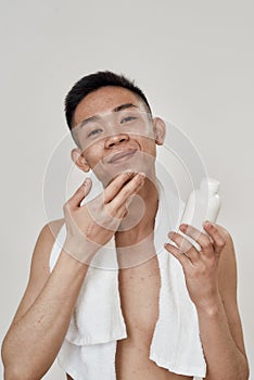 Portrait of shirtless young asian man with towel around his neck applying lotion after shaving isolated over white