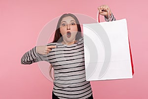 Portrait of shocked amazed woman in striped sweatshirt pointing shopping bag and looking surprised