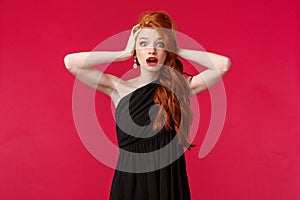 Portrait of shocked and ambushed young redhead female in elegant black dress, gasping nervously, look alarmed camera