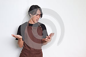 Portrait of shocked Asian barista man in brown apron looking at his mobile phone with surprised expression. Advertising concept.