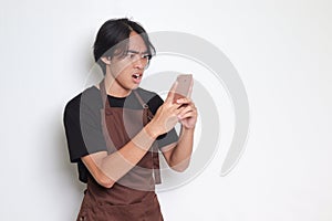 Portrait of shocked Asian barista man in brown apron looking at his mobile phone with surprised expression. Advertising concept.