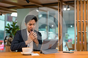 Portrait of shocked Asian businessman in coffee shop using digital tablet computer