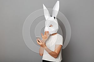 Portrait of shocked astonished anonymous woman wearing white t shirt and paper rabbit mask standing isolated over gray background