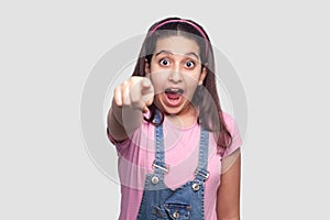 Portrait of shocked beautiful brunette young girl in casual pink t-shirt and blue overalls standing, looking and pointing at camer