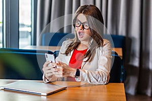 Portrait of shocked beautiful stylish brunette young woman in glasses sitting, holding mobile smart phone and looking at screen